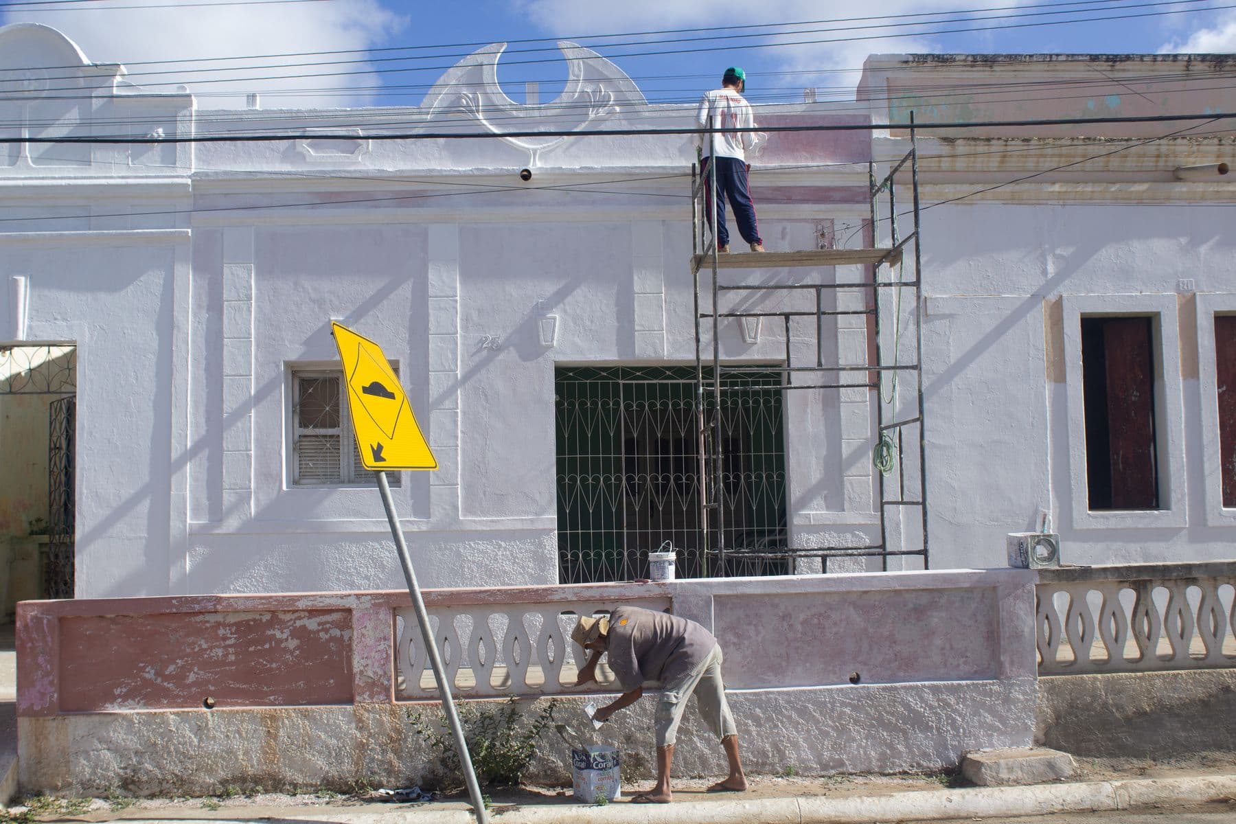 Two workers painting a white masonry facade, one on a steel scaffold and one at the base wall.
