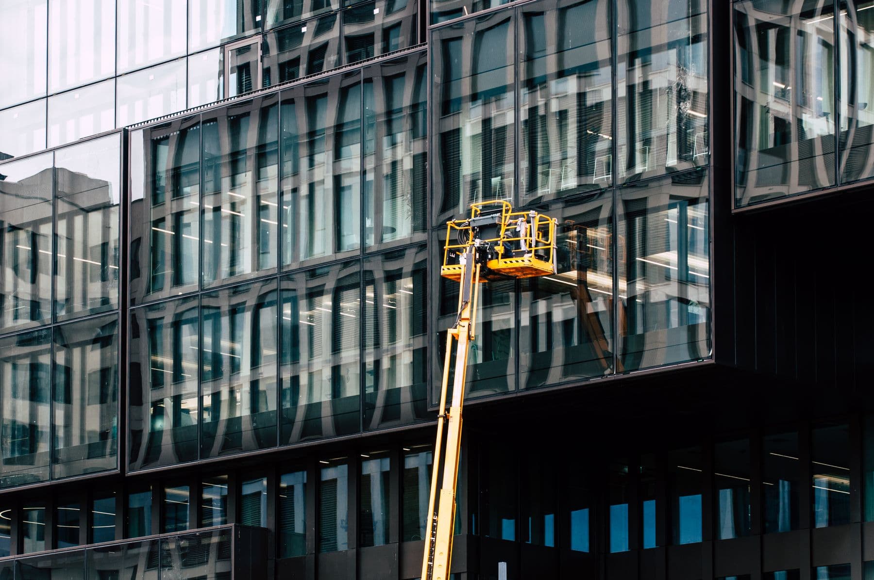 A yellow boom lift raised against the dark glass facade of a modern office building.
