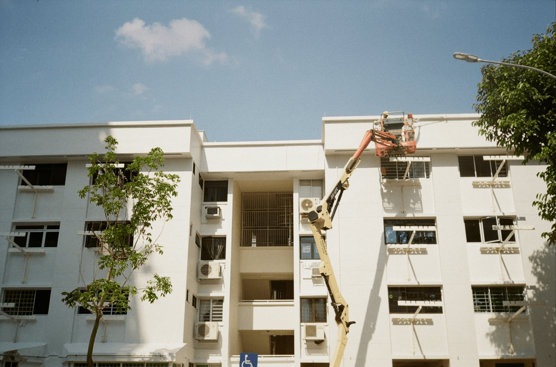 An orange boom lift working at the top floor of a white five-story apartment building.