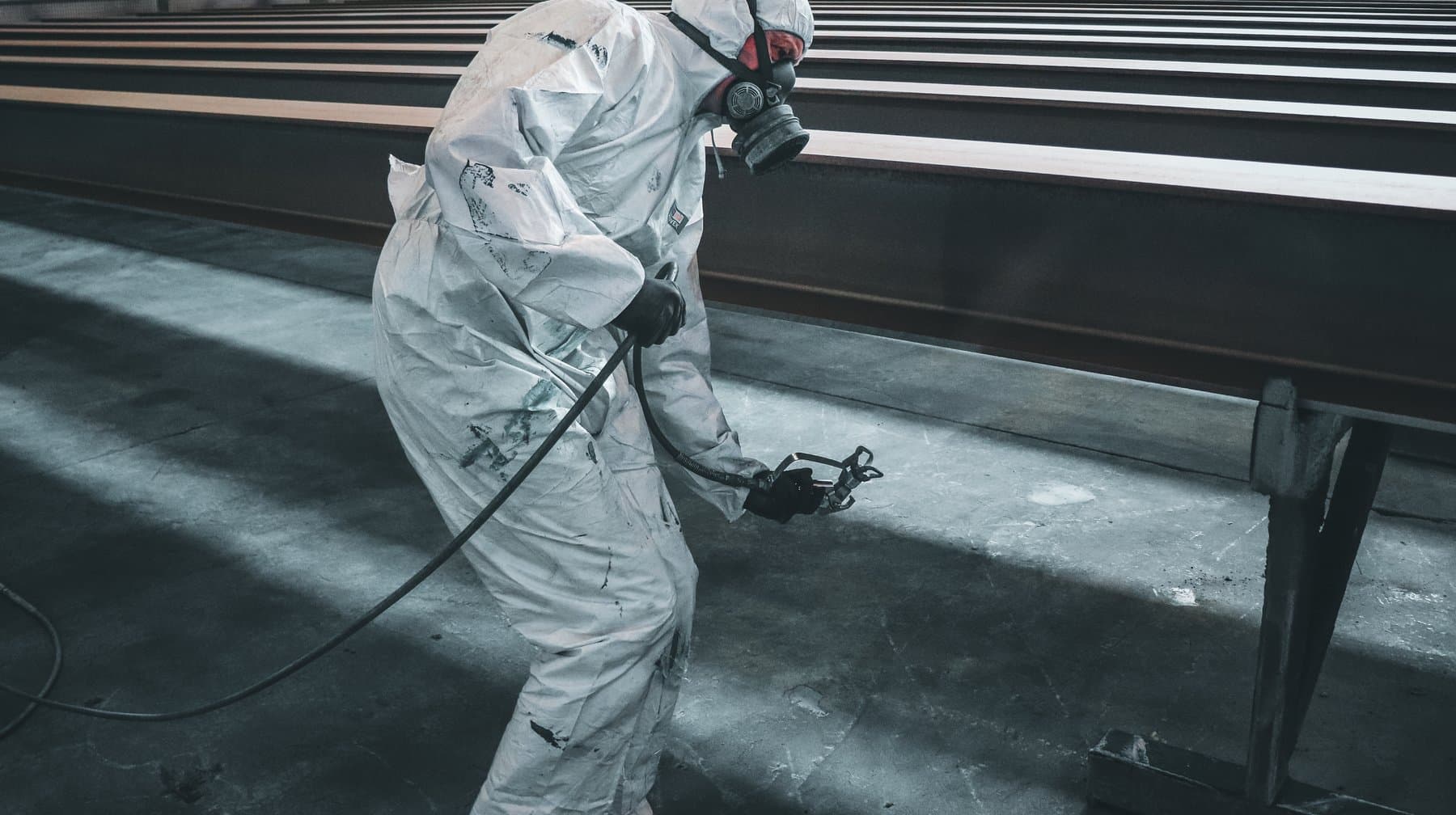 A worker in a protective suit and respirator spraying coating onto steel beams indoors.